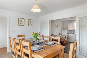 Uma sala de jantar com uma mesa de madeira posta para quatro pessoas, rodeada por cadeiras de madeira. Há um vaso com flores sobre a mesa, e cozinha visível ao fundo.
