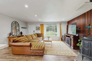 A living room with a leather sofa, a wooden TV stand, a fireplace, and a large window with curtains. - Family Favorite with Large Yard and BBQ (Fort Bragg)