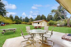 A patio with a glass-top table and four chairs, a picnic table, and a small wooden structure in the background. - Family Favorite with Large Yard and BBQ (Fort Bragg)