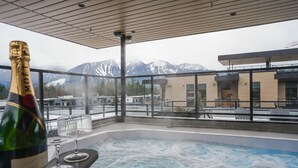 A hot tub with a mountain view, a bottle of champagne, and a modern building in the background. - Revy Retreat in Mackenzie Village (Revelstoke)