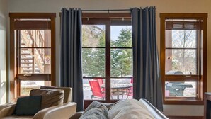 A room with a large sliding glass door leading to an outdoor area, a beige sofa with a throw blanket, and a red chair visible outside. - Aux Étoiles EMG: Ski In/Out at Soleil Express (Mont-Tremblant)