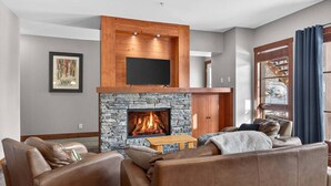 A living room with a stone fireplace, leather sofas, and a mounted TV. - Aux Étoiles EMG: Ski In/Out at Soleil Express (Mont-Tremblant)