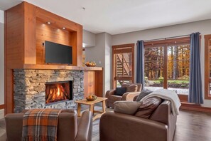 A cozy living room with a stone fireplace, leather sofas, a flat-screen TV, and a large window offering a view of trees. - Aux Étoiles EMG: Ski In/Out at Soleil Express (Mont-Tremblant)