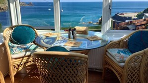Varanda com cadeiras de vime e mesa de vidro, com vista para a costa, barcos e terreno rochoso.