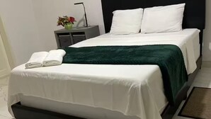Una cama bien hecha con sábanas blancas y una manta verde, un cabecero negro y una mesita de noche con un florero.