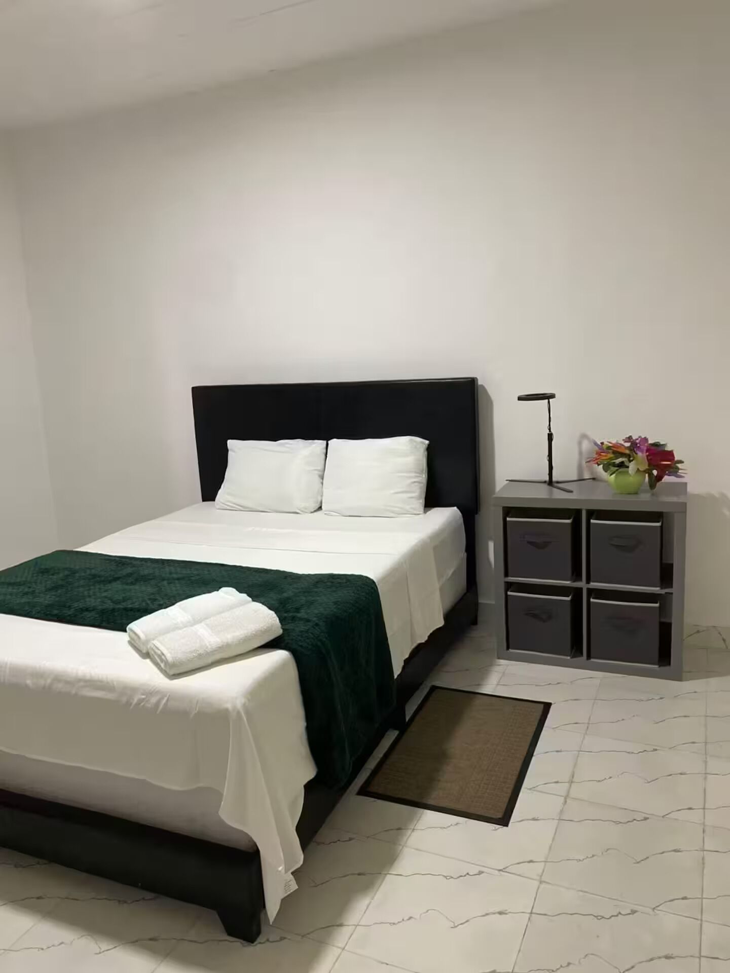 A neatly made bed with white bedding and a green throw, a bedside table with a lamp and a vase of flowers, and a storage unit with drawers.