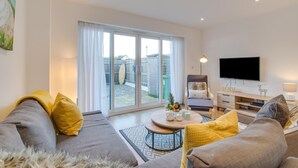 Una sala moderna con un sofá, un sillón y una mesa de centro. Hay un televisor montado en la pared y un ventanal corredero que da a un jardín.