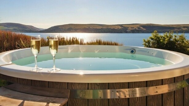 Un jacuzzi con dos copas de champán, rodeado de una terraza de madera y con vistas a un lago con colinas al fondo.