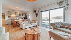 Un salón espacioso con un amplio ventanal que ofrece vistas al mar, una zona de estar acogedora con una mesa de centro redonda de madera y una cocina moderna al fondo.