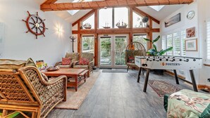 Una habitación amplia con techos altos, vigas de madera, un ventanal grande, mesa de fútbol de mesa, muebles de mimbre y un adorno de timón de barco.