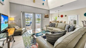 A spacious living room with a large sofa, a glass coffee table, and a television. There is a balcony with a view of the outdoors. - Poolside 5BR Escape with Pool Table & Parking (Kissimmee)