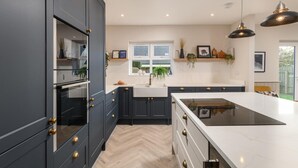Een moderne keuken met donkere kastjes, een wit aanrecht en een groot eiland. Er hangen hanglampen boven het eiland en een raam met planten op de vensterbank.