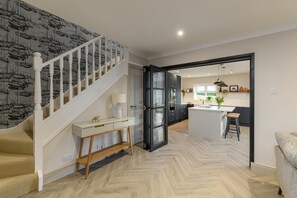 A modern kitchen with a central island, a staircase with a patterned wallpaper, and a dining area with a sofa. - 3 Bed in Bamburgh (oc-d33434) (Bamburgh)