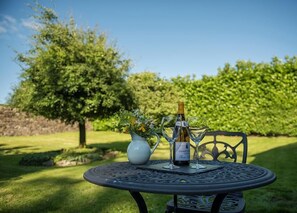 Une table ronde en métal, avec une bouteille de vin et deux verres, placée à l’extérieur, avec une haie et un arbre en arrière-plan.