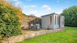 Un mur de pierre, un petit bâtiment blanc avec une porte, et une terrasse avec des chaises et une table.