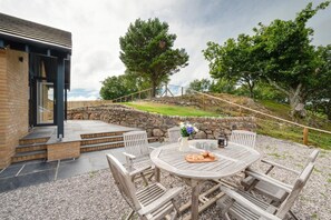 A patio with a round table and chairs, a stone wall, and a view of a grassy area with trees. - 3 Bed in Barmouth (oc-wah701) (Barmouth)