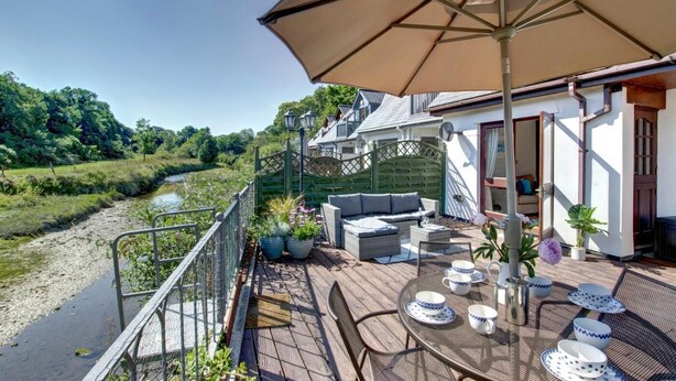 Una terraza con mobiliario exterior, una mesa dispuesta con platos y tazas, y vistas a un río y follaje.