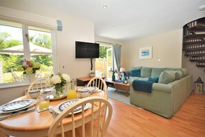 A living room with a dining table set for two, a green sofa, a flat-screen TV mounted on the wall, and a window with a view of greenery. - 2 Bed in Little Petherick (oc-p00635) (Little Petherick)