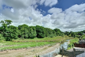 A river flowing through a forested area with a wooden bridge and a fence. - 2 Bed in Little Petherick (oc-p00635) (Little Petherick)