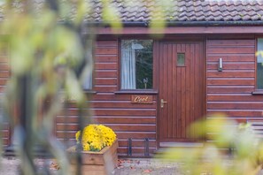 A wooden cabin with a door labeled 'Coat,' a window with a curtain, and a planter with yellow flowers. - 2 Bed in Smallburgh (oc-c33736) (Low Street)