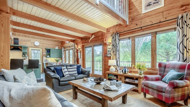 Interior acogedor de una cabaña con comedor, sofá, sillón de cuadros y techo de madera.