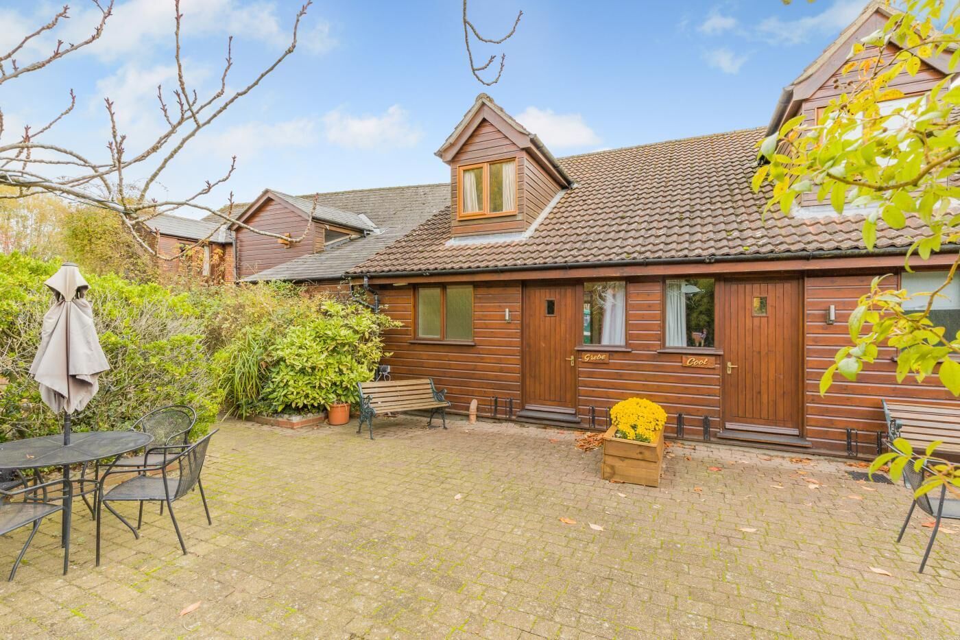 A wooden cottage with a tiled roof, a brick patio, and a small garden area with a bench and potted plants.