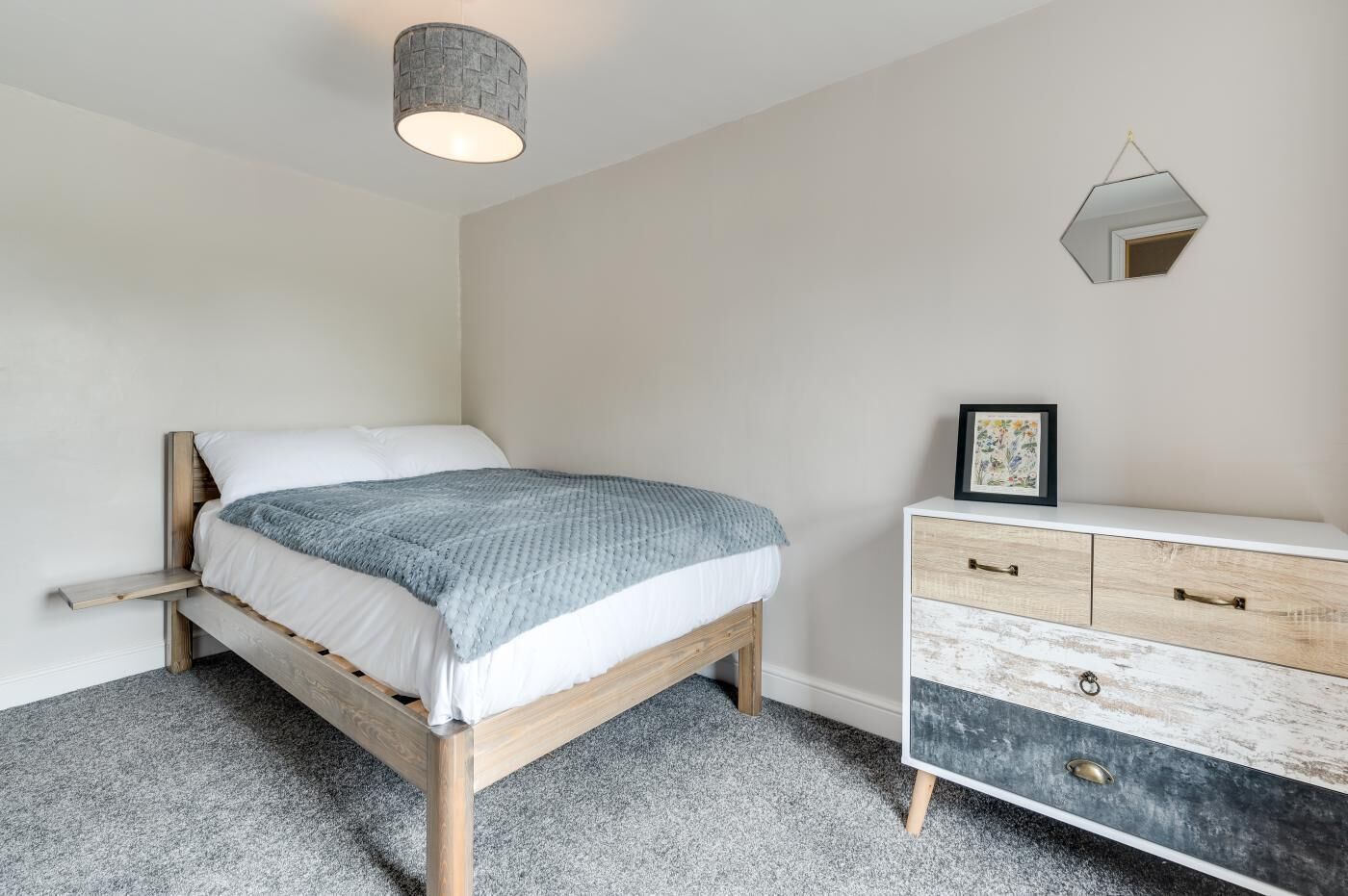 A bedroom with a wooden bed, a grey blanket, a white dresser, and a hanging light fixture.