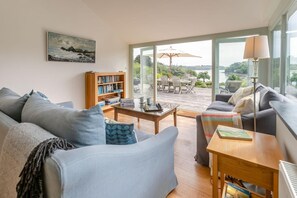 A modern living room with a sofa, coffee table, bookshelf, and a view of the outdoors. - 4 Bed in Helford (oc-hclith) (Helford)