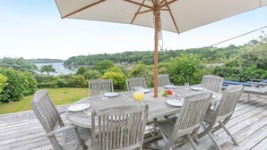 Een houten terras buiten met een ronde tafel en stoelen, een parasol en uitzicht op een meer en groen.