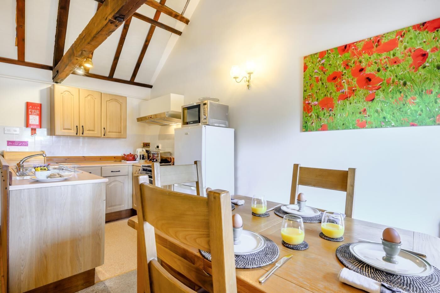 A kitchen with wooden cabinets, a dining table set for four, and a painting of red flowers on the wall.