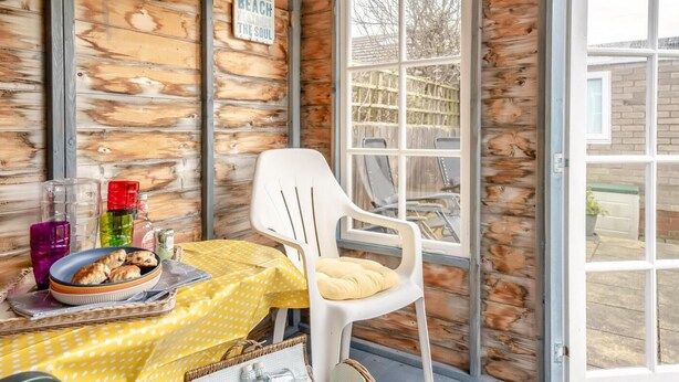 Interno di una baita rustica con parete in legno, una sedia di plastica bianca, un tavolo giallo con tovaglia a pois e una finestra che si affaccia su un giardino.