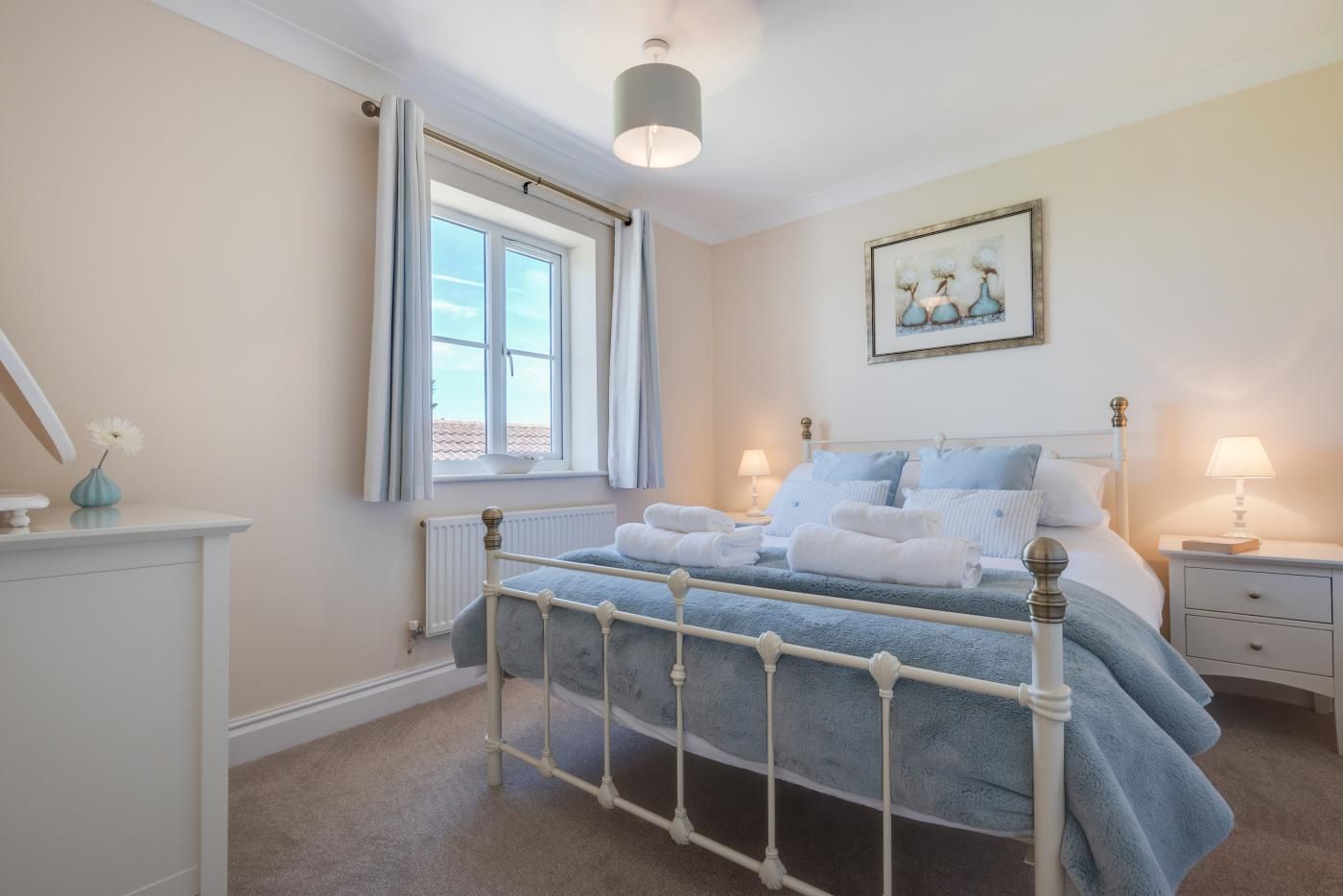 A bedroom with a bed, bedside tables, a window with curtains, a radiator, and a painting on the wall.