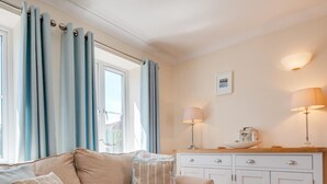 A living room with a beige sofa, two side tables with lamps, a white cabinet, and a window with blue curtains. - 3 Bed in Trimingham (oc-2290) (Trimingham)
