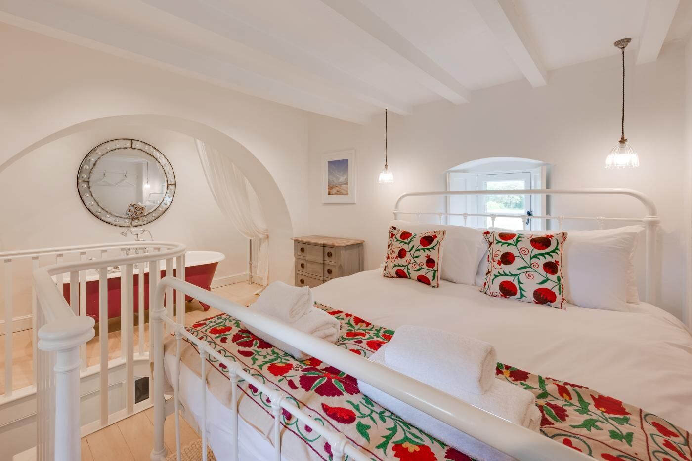 A bedroom with a white bed, floral pillows, a round mirror, a red bathtub, and a wooden nightstand.
