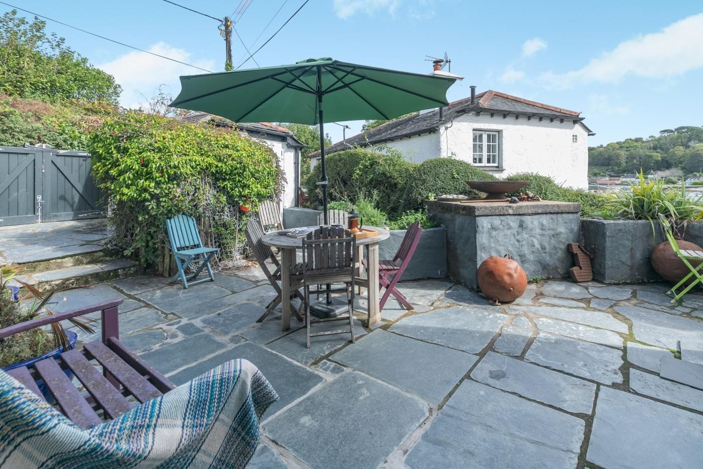 Eine Terrasse mit Steinbelag, einem Tisch mit Stühlen, einem grünen Sonnenschirm und Topfpflanzen.