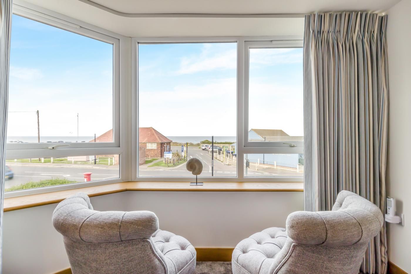 A room with a bay window overlooking a road and buildings, two upholstered armchairs, and a striped curtain.
