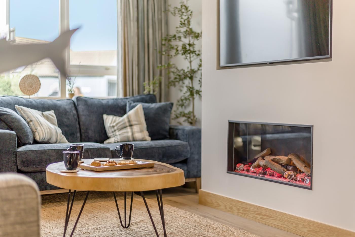 A cozy living room with a grey sofa, a wooden coffee table with cups and pastries, and a fireplace with logs.