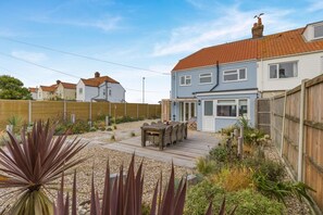 Ein zweistöckiges Haus mit einer Holzterrasse, einer Terrasse und einem Garten mit verschiedenen Pflanzen.