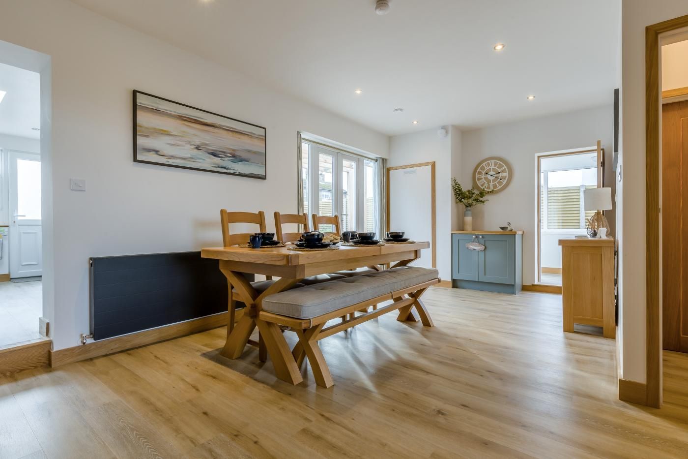 A modern kitchen with a wooden dining table, chairs, and a bench. There is a clock on the wall and a window providing natural light.