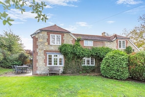 Une maison en pierre à deux étages, avec une façade en briques, un petit porche à l’avant et un jardin agrémenté d’une table et de chaises.