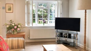 En stue med et fjernsyn på et glasbord, et træ sofabord, en sofa, et vindue med hvide skodder og en radiator.