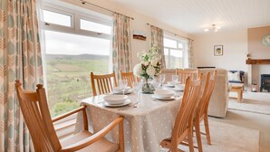 A dining room with a table set for four, wooden chairs, a vase with flowers, and a view of the countryside through large windows. - 3 Bed in Rothbury (oc-nb1976) (Rothbury)