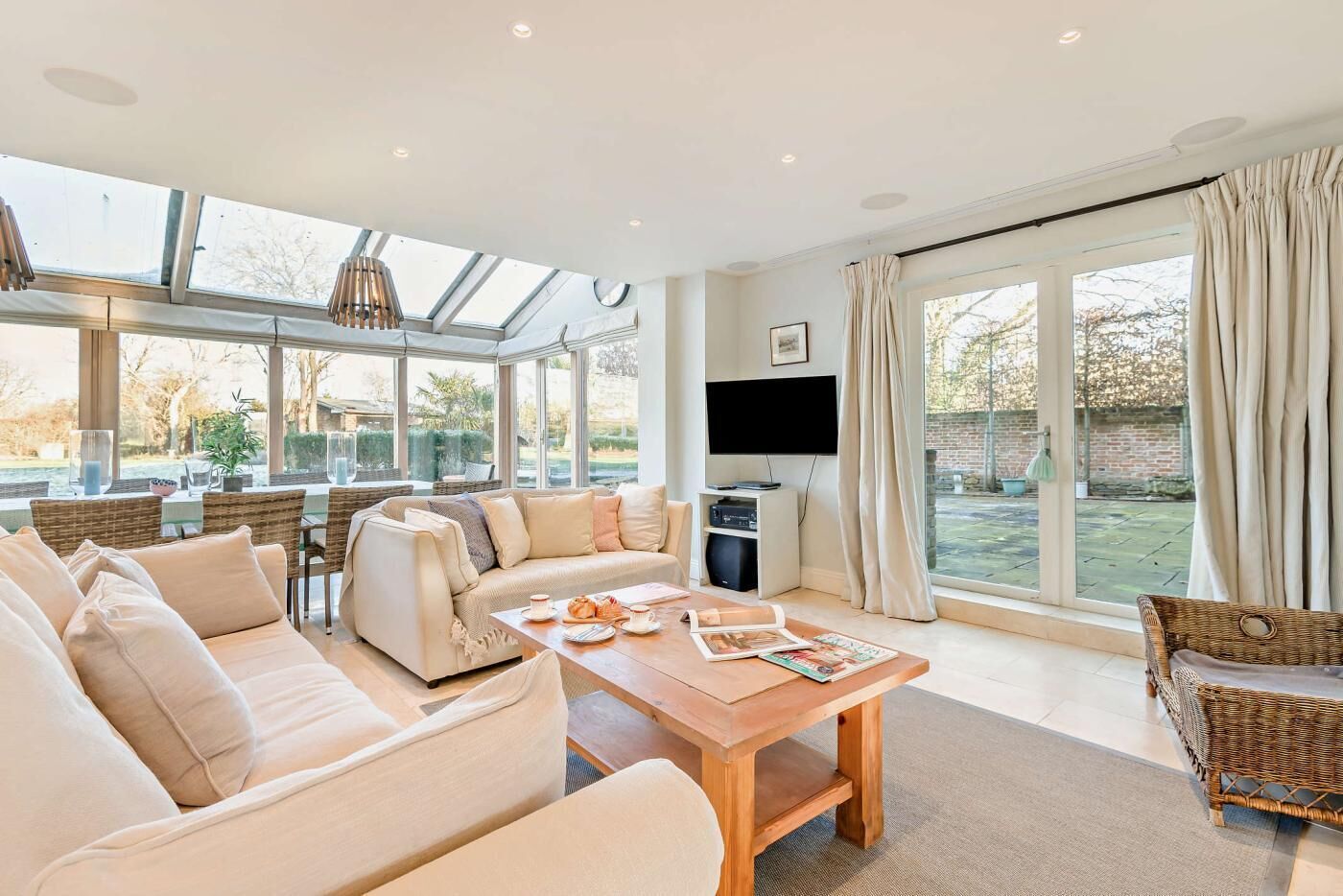 A spacious living room with a large sofa, a wooden coffee table, and a television. There are large windows providing natural light and a view of the outdoors.