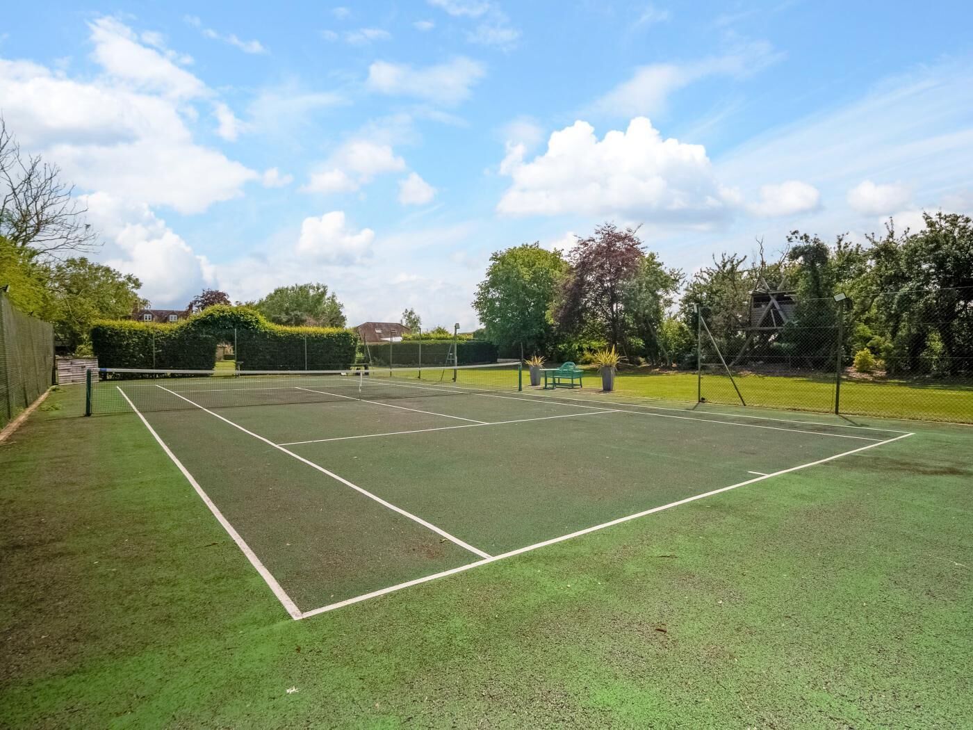 Ein Tennisplatz im Freien mit Netz, umzäunt und von Grünflächen umgeben.