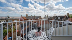 Un balcón con una mesa y sillas de metal blanco, con vistas a una zona residencial de casas con tejados rojos y un cielo azul despejado con nubes.