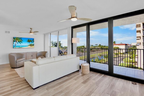 Una sala de estar moderna con un sofá blanco, un sillón color beige y un ventilador de techo. Amplias ventanas ofrecen vistas a un área exterior ajardinada.