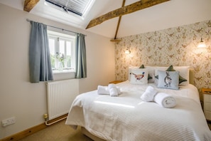 A bedroom with a bed, pillows, a window with curtains, a radiator, and a wall with floral wallpaper.