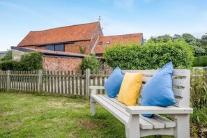 Eine Holzbank mit Kissen, ein Backsteinhaus mit Ziegeldach und ein Holzzaun in einem Garten.