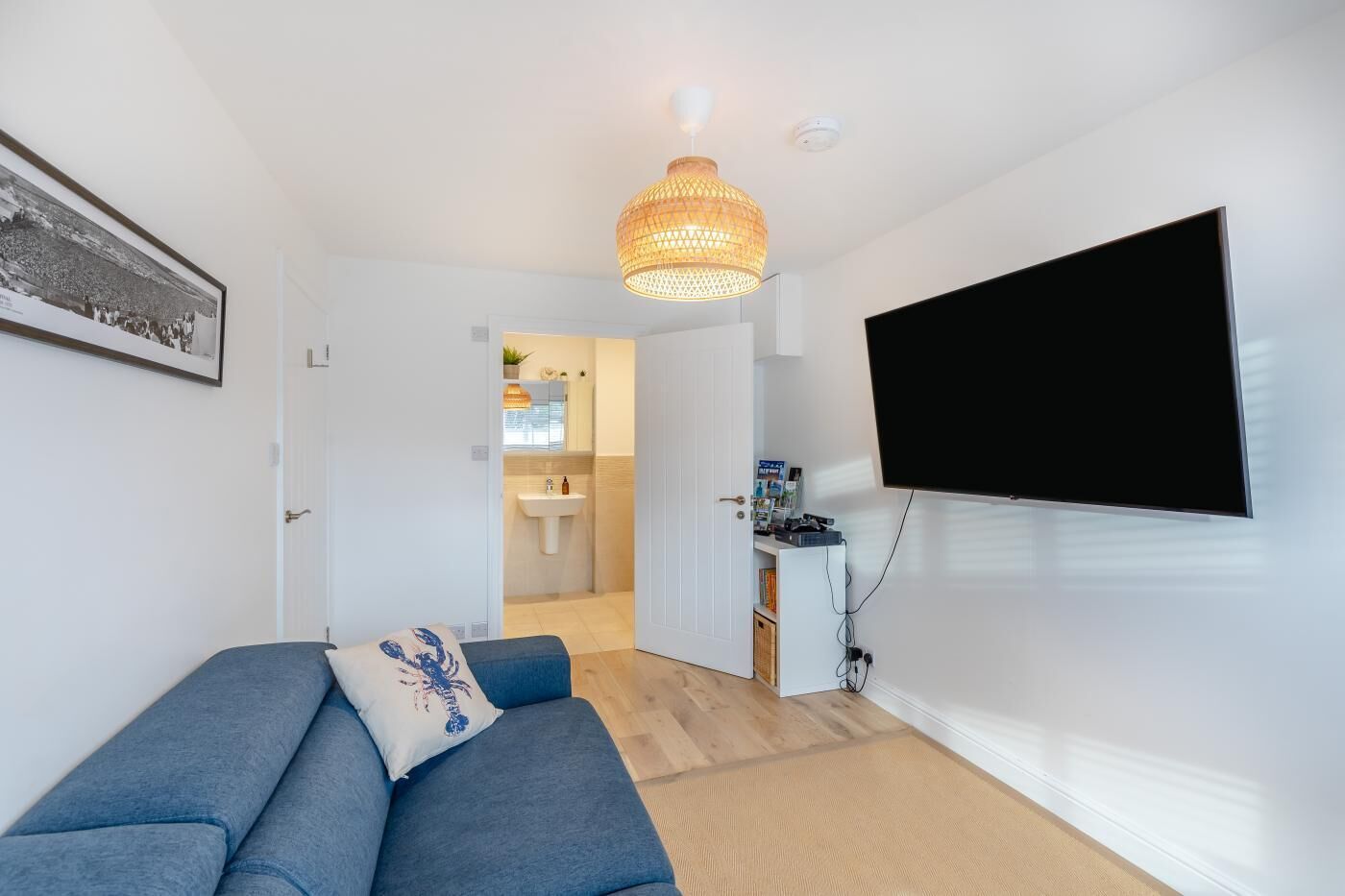 A modern living room with a sofa, a mounted TV, and a kitchen area visible through an open door.