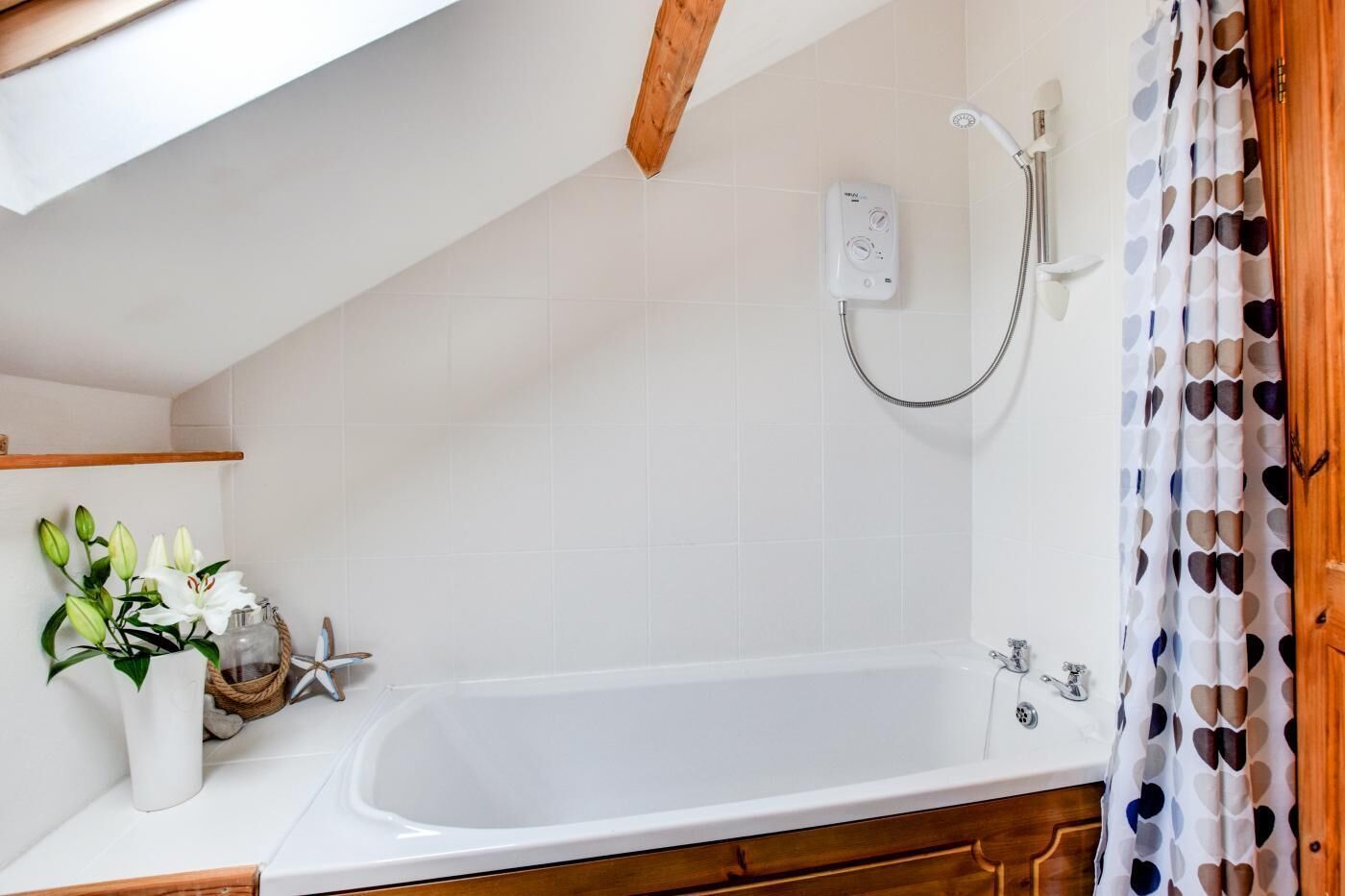 Un baño con una bañera blanca, ducha y un perchero. Se aprecia una viga de madera en el techo y un estante blanco con un jarrón de flores.